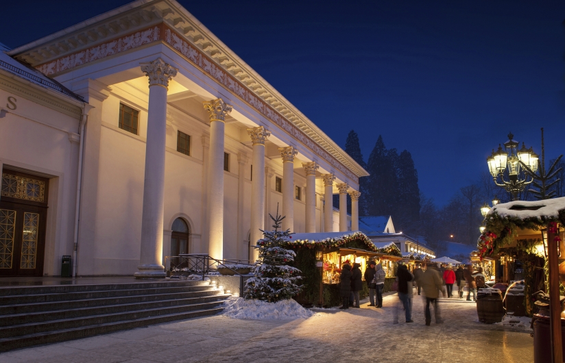 Fotografie zur Zeit der blauen Stunde im Winter. Auf der linken Seite erstreckt sich die weiße, klassizistische Fassade des Kurhauses Baden-Baden. Sie besitzt eine monumentale Vorhalle, die von hohen, von unten her angestrahlten Säulen mit korinthischen Kapitellen getragen wird. Eine breite Steintreppe führt zum Eingang hinauf. Der Boden und die Dächer sind mit einer dünnen Schneeschicht bedeckt. Vor dem Gebäude verläuft eine weihnachtliche Marktstraße. Auf der rechten Seite reihen sich hölzerne, festlich beleuchtete Marktbuden aneinander. Die Buden sind mit Tannengrün dekoriert, ihre Dächer tragen Schnee. Mehrere Passanten in winterlicher Kleidung schlendern an den Ständen entlang. Im Vordergrund steht ein kleiner, schneebedeckter Weihnachtsbaum. Eine historische, dreiflammige Straßenlaterne aus Gusseisen auf der rechten Seite wirft warmes Licht auf die Szene. Im Hintergrund ragen dunkle Tannenbäume in den tiefblauen Abendhimmel.