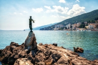 Küste mit Statue auf Felsen, Meer und Hügel im Hintergrund.