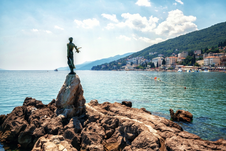 Küste mit Statue auf Felsen, Meer und Hügel im Hintergrund.