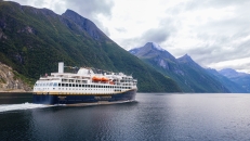 Eine Totalaufnahme eines großen, modernen Passagierschiffs, das von links nach rechts durch ein ruhiges, dunkelblaues Gewässer fährt und eine weiße Heckwelle hinterlässt. Das Schiff hat einen dunkelblauen Rumpf mit einem markanten goldenen Längsstreifen. Auf der Bordwand steht in goldenen Großbuchstaben „HAVILA KYSTRUTEN“. Darüber befinden sich mehrere weiße Decksetagen mit zahlreichen Fenstern und Kabinenbalkonen sowie orangefarbene Rettungsboote. Auf dem oberen Deck ist eine bunte Flaggenkette gespannt. Am Heck ist der Name „HAVILA POLLUX“ zu lesen. Das Schiff befindet sich in einem Fjord, der von steilen, massiven Bergen umgeben ist. Die Berghänge sind im unteren Bereich dicht mit grünem Wald bewachsen, während die oberen Gipfel schroff und felsig sind. Der Himmel ist weitgehend von einer dichten, hellgrauen Wolkendecke überzogen, durch die vereinzelt hellere Lichtblicke dringen.