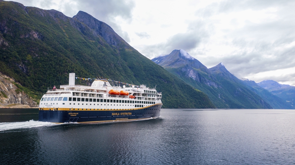 Eine Totalaufnahme eines großen, modernen Passagierschiffs, das von links nach rechts durch ein ruhiges, dunkelblaues Gewässer fährt und eine weiße Heckwelle hinterlässt. Das Schiff hat einen dunkelblauen Rumpf mit einem markanten goldenen Längsstreifen. Auf der Bordwand steht in goldenen Großbuchstaben „HAVILA KYSTRUTEN“. Darüber befinden sich mehrere weiße Decksetagen mit zahlreichen Fenstern und Kabinenbalkonen sowie orangefarbene Rettungsboote. Auf dem oberen Deck ist eine bunte Flaggenkette gespannt. Am Heck ist der Name „HAVILA POLLUX“ zu lesen. Das Schiff befindet sich in einem Fjord, der von steilen, massiven Bergen umgeben ist. Die Berghänge sind im unteren Bereich dicht mit grünem Wald bewachsen, während die oberen Gipfel schroff und felsig sind. Der Himmel ist weitgehend von einer dichten, hellgrauen Wolkendecke überzogen, durch die vereinzelt hellere Lichtblicke dringen.