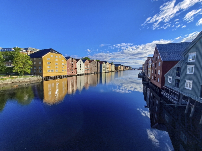 Eine Weitwinkelaufnahme von einer Brücke herab auf den Fluss Nidelva, der sich geradlinig in die Ferne erstreckt. Das ruhige, tiefblaue Wasser reflektiert die Uferbebauung und die weißen Wolken des Himmels. Zu beiden Seiten des Flusses reihen sich historische, mehrstöckige Holzlagerhäuser auf Holzpfählen aneinander. Die Häuser auf der linken Seite sind in leuchtenden Farben wie Gelb, Rot und Weiß gestrichen, während auf der rechten Seite dunklere rote, braune und graue Holzhäuser dominieren. Am linken Ufer verläuft zudem eine Uferpromenade mit einer grünen Wiese und Bäumen. Der Himmel ist kräftig blau mit lockeren, weißen Schleierwolken.