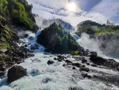 Dramatische Weitwinkel-Außenaufnahme des Låtefossen-Wasserfalls unter einem hellen, von Schleierwolken bedeckten Himmel. Im gesamten Vorder- und Mittelgrund bahnen sich wilde, schäumend weiße Wassermassen reißend ihren Weg durch ein Bett aus unzähligen großen, dunklen Felsbrocken und Steinen.Im Hintergrund stürzt der Wasserfall in zwei gewaltigen Kaskaden von den oberen Berghängen herab. Die beiden mächtigen Ströme fließen links und rechts an einem zentralen, steilen Felskegel vorbei, der dicht mit grünen Nadelbäumen bewachsen ist. Aus den herabstürzenden Fluten steigt eine dichte, weiße Gischtwolke auf, die besonders die rechte Bildhälfte und die bewaldeten Hänge im Hintergrund in einen diffusen Nebel hüllt. Die Sonne steht hoch über der Szenerie hinter dünnen Wolken und lässt das Wasser hell aufleuchten.