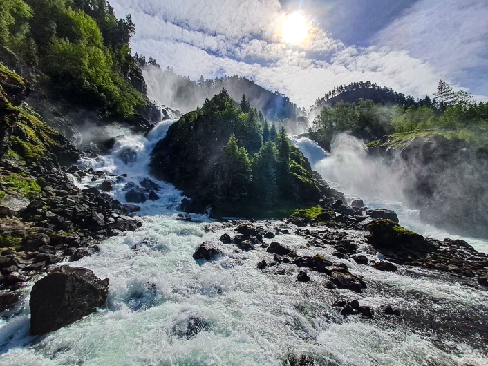 Dramatische Weitwinkel-Außenaufnahme des Låtefossen-Wasserfalls unter einem hellen, von Schleierwolken bedeckten Himmel. Im gesamten Vorder- und Mittelgrund bahnen sich wilde, schäumend weiße Wassermassen reißend ihren Weg durch ein Bett aus unzähligen großen, dunklen Felsbrocken und Steinen.Im Hintergrund stürzt der Wasserfall in zwei gewaltigen Kaskaden von den oberen Berghängen herab. Die beiden mächtigen Ströme fließen links und rechts an einem zentralen, steilen Felskegel vorbei, der dicht mit grünen Nadelbäumen bewachsen ist. Aus den herabstürzenden Fluten steigt eine dichte, weiße Gischtwolke auf, die besonders die rechte Bildhälfte und die bewaldeten Hänge im Hintergrund in einen diffusen Nebel hüllt. Die Sonne steht hoch über der Szenerie hinter dünnen Wolken und lässt das Wasser hell aufleuchten.