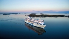 Vogelperspektive auf ein großes, weißes Kreuzfahrtschiff mit blauem Aufdruck „SILJA LINE“ an der Bordwand. Das Schiff fährt von links nach rechts durch eine ruhige, dunkelblaue Meeresoberfläche und hinterlässt eine sanfte Heckwelle. Im Hintergrund erstrecken sich flache, bewaldete Schäreninseln unter einem weiten Himmel, der am Horizont in zarten Rosa- und Pastelltönen des Abendlichts schimmert. Das Schiff und die Inseln spiegeln sich klar auf der Wasseroberfläche.