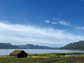 Eine horizontale Landschaftsaufnahme einer skandinavischen Fjordlandschaft bei sonnigem Tageslicht. Das untere linke Viertel des Bildes wird von einer kleinen, rechteckigen, dunkelbraunen Holzhütte eingenommen, deren Satteldach dicht mit üppigem, grünem Gras bewachsen ist. Die Hütte steht auf einer weitläufigen, grünen Wiese mit vereinzelt gelb blühenden Wildblumen, die den gesamten Vordergrund einnimmt. Im rechten Mittelgrund erstreckt sich eine schmale, grüne Landzunge mit einer Schilf- und Graszone, an deren Rand eine kleine Bucht liegt. Auf der Wiese im Mittelgrund halten sich vereinzelt kleine Personensilhouetten auf. Dahinter öffnet sich der Blick auf die weite, ruhige und hellblaue Wasseroberfläche des Fjords. Am gegenüberliegenden Ufer im Hintergrund zieht sich eine langgestreckte, massive Gebirgskette durch das Bild, deren steile, grüne und graue Flanken sanft ins Wasser abfallen. Auf der rechten Seite ragen schroffe, felsige Bergspitzen empor. Den oberen Teil des Bildes dominiert ein weiter, intensiv blauer Himmel, der im mittleren Bereich von dünnen, weißen Schleierwolken durchzogen ist.