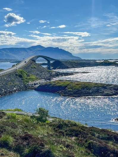 Eine vertikale Landschaftsaufnahme einer Küstenregion mit einer modernen Brücke bei sonnigem Tageslicht. Der unmittelbare Vordergrund wird von einer hügeligen, dunkelgrünen Böschung eingenommen, die mit kurzem Gras, Moos und Heidekraut bewachsen ist. Am Fuß des Hangs ist ein kleiner Ausschnitt des hellgrauen Gitterrost-Wanderwegs mit Edelstahlgeländer zu erkennen. Dahinter erstreckt sich eine weite, dunkelblaue Meeresoberfläche, auf der das Sonnenlicht intensiv silbern glitzert. Mehrere flache, dunkelgraue und teils grün bewachsene Schäreninseln ragen aus dem Wasser. Auf der linken Seite zieht sich die aufgeschüttete Rampe der Atlantikstraße über die Felsen nach hinten. Auf der zweispurigen Asphaltstraße fährt ein einzelnes, dunkles Auto auf die Storseisundet-Brücke zu. Die Brücke überspannt den Meeresarm mit einer hohen, eleganten Betonbogenkonstruktion, die im Mittelgrund zu einer markanten Kurve ansteigt. Den Hintergrund bildet eine massive Gebirgskette im blauen Dunst unter einem weiten, hellblauen Himmel, der von dünnen, langgestreckten weißen Wolkenbändern durchzogen wird.
