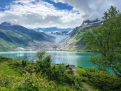 Das im Querformat aufgenommene Foto zeigt eine weite alpine Fjord- und Gletscherlandschaft unter einem teils bewölkten Himmel in Norwegen. Im unteren Bilddrittel erstreckt sich ein hügeliger Vordergrund, der dicht mit grünen Büschen, Sträuchern und vereinzelten, jungen Birken bewachsen ist. Auf einer kleinen, unbewachsenen Kuppe inmitten dieser Vegetation stehen im rechten unteren Bildzentrum zwei Personen von hinten fotografiert. Sie blicken nach vorne auf einen großen, spiegelnden Gletschersee, der das mittlere Bilddrittel einnimmt. Das ruhige Wasser besitzt eine charakteristische, milchig-türkise Färbung. Im Hintergrund des Sees ragt eine gewaltige Bergkette empor. Zwischen den steilen, kargen Felswänden der Berge erstreckt sich im Zentrum eine mächtige, breite Gletscherzunge, die sanft bis kurz vor das Seeufer hinabfällt. Das Eis leuchtet weiß-blau. Der Himmel darüber ist dynamisch bewölkt: Große, weiße, von der Sonne beschienene Wolkenbänke wechseln sich mit grauen Regenwolken ab, die teilweise die Bergspitzen einhüllen. Das Licht ist hell und wirft weiche Spiegelungen auf die Wasseroberfläche.