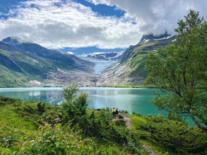 Das im Querformat aufgenommene Foto zeigt eine weite alpine Fjord- und Gletscherlandschaft unter einem teils bewölkten Himmel in Norwegen. Im unteren Bilddrittel erstreckt sich ein hügeliger Vordergrund, der dicht mit grünen Büschen, Sträuchern und vereinzelten, jungen Birken bewachsen ist. Auf einer kleinen, unbewachsenen Kuppe inmitten dieser Vegetation stehen im rechten unteren Bildzentrum zwei Personen von hinten fotografiert. Sie blicken nach vorne auf einen großen, spiegelnden Gletschersee, der das mittlere Bilddrittel einnimmt. Das ruhige Wasser besitzt eine charakteristische, milchig-türkise Färbung. Im Hintergrund des Sees ragt eine gewaltige Bergkette empor. Zwischen den steilen, kargen Felswänden der Berge erstreckt sich im Zentrum eine mächtige, breite Gletscherzunge, die sanft bis kurz vor das Seeufer hinabfällt. Das Eis leuchtet weiß-blau. Der Himmel darüber ist dynamisch bewölkt: Große, weiße, von der Sonne beschienene Wolkenbänke wechseln sich mit grauen Regenwolken ab, die teilweise die Bergspitzen einhüllen. Das Licht ist hell und wirft weiche Spiegelungen auf die Wasseroberfläche.