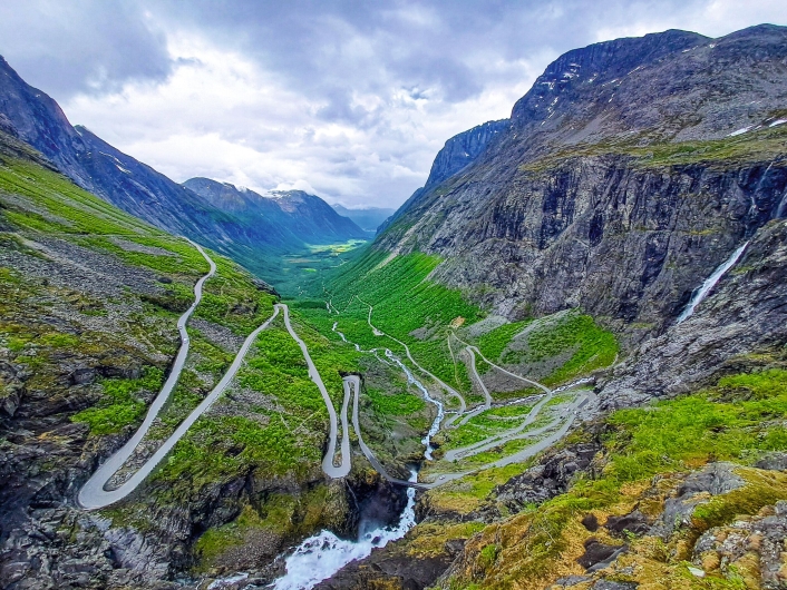 Blick aus großer Höhe in ein tiefes, grünes Gletschertal, das von steilen, massiven Bergen eingerahmt wird. An den Hängen windet sich die schmale Passstraße Trollstigen in engen, s-förmigen Haarnadelkurven hinab. Am Fuße der Felswände fließt ein reißender Gebirgsfluss, der im Vordergrund als weißer Wasserfall unter einer kleinen Brücke hindurchstürzt. Die Berghänge sind im oberen Teil karg und felsig, weiter unten mit sattgrüner Vegetation bewachsen. Der Himmel über dem Tal ist von einer dichten, dramatischen Decke aus grauen und weißen Wolken überzogen.