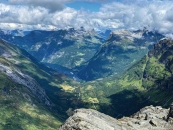 Weitwinkel-Fotografie im Querformat aus einer extrem hohen Bergperspektive. Im untersten Bilddrittel ragen schroffe, graue und von Flechten besiedelte Felsformationen des Dalsnibba-Gipfels in den Vordergrund. Dahinter blickt man in ein gewaltiges, tief eingeschnittenes Gletschertal mit saftig grünen Hängen, kleinen Waldflecken und hellen landwirtschaftlichen Flächen im Talboden, durch die sich eine schmale Straße schlängelt. Am fernen Ende des Tals, im linken mittleren Bildbereich, schmiegt sich das dunkelblaue Wasser des Geirangerfjords in die Landschaft. Auf dem Fjord liegen gut erkennbar zwei große, weiße Kreuzfahrtschiffe vor Anker. Das Tal wird von riesigen, steilen Bergen flankiert, deren schroffe Flanken teils im Schatten von Wolken liegen. Die oberen Gipfel der massiven Gebirgskette im Hintergrund sind teilweise schneebedeckt und werden von einer dichten, schweren Schicht aus weißen und grauen Wolken eingehüllt, zwischen denen kleine Abschnitte eines hellblauen Himmels sichtbar sind.