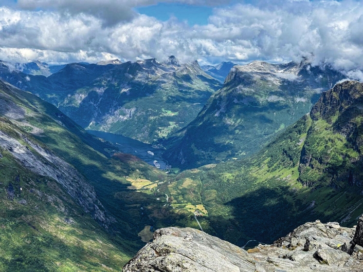 Weitwinkel-Fotografie im Querformat aus einer extrem hohen Bergperspektive. Im untersten Bilddrittel ragen schroffe, graue und von Flechten besiedelte Felsformationen des Dalsnibba-Gipfels in den Vordergrund. Dahinter blickt man in ein gewaltiges, tief eingeschnittenes Gletschertal mit saftig grünen Hängen, kleinen Waldflecken und hellen landwirtschaftlichen Flächen im Talboden, durch die sich eine schmale Straße schlängelt. Am fernen Ende des Tals, im linken mittleren Bildbereich, schmiegt sich das dunkelblaue Wasser des Geirangerfjords in die Landschaft. Auf dem Fjord liegen gut erkennbar zwei große, weiße Kreuzfahrtschiffe vor Anker. Das Tal wird von riesigen, steilen Bergen flankiert, deren schroffe Flanken teils im Schatten von Wolken liegen. Die oberen Gipfel der massiven Gebirgskette im Hintergrund sind teilweise schneebedeckt und werden von einer dichten, schweren Schicht aus weißen und grauen Wolken eingehüllt, zwischen denen kleine Abschnitte eines hellblauen Himmels sichtbar sind.