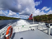 Vom grauen Außendeck am Heck des schnell fahrenden Schiffes richtet sich der Blick nach hinten in eine schmale Meerenge. Im rechten Vordergrund weht eine rote norwegische Flagge mit blau-weißem Kreuz an einem weißen Mast. Eine weiße Metallreling begrenzt das Deck, an deren linkem Rand ein orangefarbener Rettungsring angebracht ist. Zentral zieht sich eine breite, wild aufgewühlte Bahn aus strahlend weißem Schaum durch das dunkelblaue Wasser. Die Meerenge wird links von einem dicht bewaldeten, flachen Grashügel begrenzt. Auf der rechten Seite steigt das Ufer als felsiger, grüner Hang an, auf dessen Kuppe im Hintergrund mehrere weiße Wohnhäuser stehen. Der weite Himmel ist von großen, bauschigen weißen und dunkelgrauen Wolkenbänken bedeckt, zwischen denen strahlend blaue Lücken zu sehen sind.