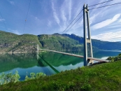 Weitwinkel-Außenaufnahme einer großen Hängebrücke an einem sonnigen Tag unter einem hellblauen Himmel mit feinen, federartigen Schleierwolken. Im rechten Vordergrund ragt ein mächtiger, grauer Betonpylon steil in die Höhe, von dem dicke Tragkabel schräg nach unten über den Fjord führen. Die Brückenfahrbahn verläuft von rechts vorne flach ansteigend über das Wasser und führt am gegenüberliegenden Ufer in einen Tunnel im Berghang, wo ein kleinerer, weißer Pylon zu sehen ist.Das Wasser des Fjords schimmert in einem tiefen Türkisgrün und spiegelt die umliegende Landschaft wider. Im Hintergrund erhebt sich eine massive, steile Bergkette, die dicht mit grünen Wäldern bewachsen ist und nach oben hin in felsige, graue Gipfel übergeht. Der Vordergrund wird von einer erhöhten, grasbewachsenen Böschung eingenommen, auf der mehrere schlanke Nadelbäume und Kiefern stehen, deren Kronen in das Bild ragen. Das Sonnenlicht erhellt die gesamte Szenerie von vorne links.