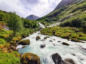 Ein breiter, wild reißender Wildbach mit milchig-weißem, stark schäumendem Wasser fließt aus der Bildmitte nach vorne auf den Betrachter zu. Im Flussbett und an den Ufern liegen zahlreiche große, abgerundete Felsbrocken, die dicht mit grünem und gelblichem Moos bewachsen sind. Das rechte Ufer steigt als hügelige, saftig grüne Gras- und Mooslandschaft an. Im Hintergrund blickt man auf einen großen, stufenförmigen Wasserfall, der dichte Gischt- und Nebelschwaden in die Luft wirbelt. Über den oberen Bereich des Wasserfalls spannt sich eine schmale, dunkle Brücke. Die umliegende Landschaft ist von steilen, dicht mit grünen Laubbäumen bewachsenen Berghängen geprägt. Im fernen Hintergrund ragt eine kahle, graue Bergnase unter einem hellen, bewölkten Sommerhimmel empor.