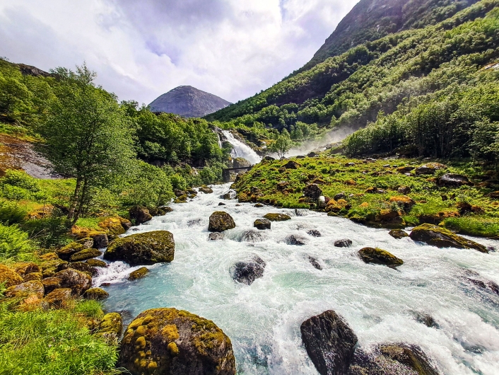 Ein breiter, wild reißender Wildbach mit milchig-weißem, stark schäumendem Wasser fließt aus der Bildmitte nach vorne auf den Betrachter zu. Im Flussbett und an den Ufern liegen zahlreiche große, abgerundete Felsbrocken, die dicht mit grünem und gelblichem Moos bewachsen sind. Das rechte Ufer steigt als hügelige, saftig grüne Gras- und Mooslandschaft an. Im Hintergrund blickt man auf einen großen, stufenförmigen Wasserfall, der dichte Gischt- und Nebelschwaden in die Luft wirbelt. Über den oberen Bereich des Wasserfalls spannt sich eine schmale, dunkle Brücke. Die umliegende Landschaft ist von steilen, dicht mit grünen Laubbäumen bewachsenen Berghängen geprägt. Im fernen Hintergrund ragt eine kahle, graue Bergnase unter einem hellen, bewölkten Sommerhimmel empor.