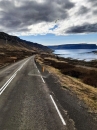 Eine vertikale Landschaftsaufnahme aus der Fahrerperspektive auf eine asphaltierte Küstenstraße unter einem stark bewölkten Himmel mit grauen Wolkenformationen und vereinzelten blauen Lücken. Die zweispurige Straße im Vordergrund ist mit einer doppelten durchgezogenen weißen Linie in der Mitte und einer gestrichelten weißen Linie am rechten Fahrbahnrand markiert. Sie verläuft leicht geschwungen in die Bildmitte. Am rechten Straßenrand steht ein einzelner oranger Markierungspfosten.Auf der linken Seite steigen direkt hinter dem Asphalt dunkle, felsige und von Geröll geprägte Berghänge steil an. Die kargen Hänge weisen flache Zonen mit trockenem, hellbraunem Grasbewuchs auf. Auf der rechten Seite fällt das Gelände sanft zu einem weiten, blassblauen Fjord ab. Am flachen Ufersaum ist die dunkle Gezeitenzone erkennbar. Den Hintergrund hinter dem Fjordwasser bildet eine massive, langgezogene Halbinsel mit flachen Tafelbergen, die im rechten Bildhintergrund steil in das Meer abbricht. Das Foto wurde durch eine Glasscheibe aufgenommen, auf der im oberen Bereich leichte Spiegelungen sichtbar sind.