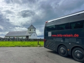 Die rechte Bildhälfte wird von der Seiten- und Heckansicht eines großen, modernen, schwarzen Reisebusses eingenommen, auf dessen Karosserie die rote Webadresse „www.blitz-reisen.de“ aufgedruckt ist. Der Bus parkt auf einer grauen Asphaltfläche. Im linken Hintergrund erstreckt sich eine niedrige, saftig grüne Wiese, auf der die weiße Sankt-Olav-Kirche steht. Die Kirche hat ein hellgraues Blechdach und einen angebauten, quadratischen Turm mit Pyramidendach und einem kleinen Kreuz an der Spitze. Das Kirchengrundstück ist im Vordergrund durch eine dicke, aus unregelmäßigen grauen Natursteinen aufgeschichtete Trockenmauer vom Parkplatz getrennt, an der im Hintergrund eine kleine Menschengruppe entlanggeht. Der Himmel ist von einer dichten, schweren Wolkendecke überzogen.