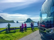 Eine horizontale Außenaufnahme bei Tageslicht unter einem leicht bewölkten, blauen Himmel. Die rechte Bildhälfte wird von der dunkelgrünen bis schwarzen Front eines modernen Setra-Reisebusses eingenommen, der am Rand einer asphaltierten Straße parkt. Links daneben stehen auf einer grünen Wiese abseits der Leitplanke fünf ältere Touristen, die mit ihren Smartphones die Landschaft fotografieren. Ein weiterer Mann sitzt links auf einer Holzbank. Im Hintergrund erstreckt sich das tiefblaue Meer, aus dem im Zentrum die spitze, pyramidenartige Silhouette der Insel Tindhólmur sowie links davon zwei markante, vertikale Felsnadeln emporragen. Am linken Bildrand ist der grüne Hang einer Küstenklippe zu sehen.