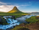 Weitwinkel-Naturaufnahme in Island bei Tageslicht. Im linken Vordergrund stürzt ein kleiner, mehrstufiger Wasserfall über moosbewachsene, dunkle Felsen. Das schäumende, weiße Wasser fließt in einem weiten Bogen als blauer Fluss durch eine saftig grüne, hügelige Landschaft nach rechts hinten weg. Im Bildzentrum erhebt sich im Hintergrund der markante, pyramiden- oder kegelförmige Berg Kirkjufell, dessen linke Flanke von der tief stehenden Sonne hellgrün erleuchtet wird. Rechts im Hintergrund ist ein ruhiger Fjord zu sehen, der von flachen Bergen gesäumt wird. Der Himmel ist strahlend blau mit einigen lockeren, weißen Schleierwolken. Die Lichtstimmung ist klar und spätnachmittags- oder morgendlich warm.