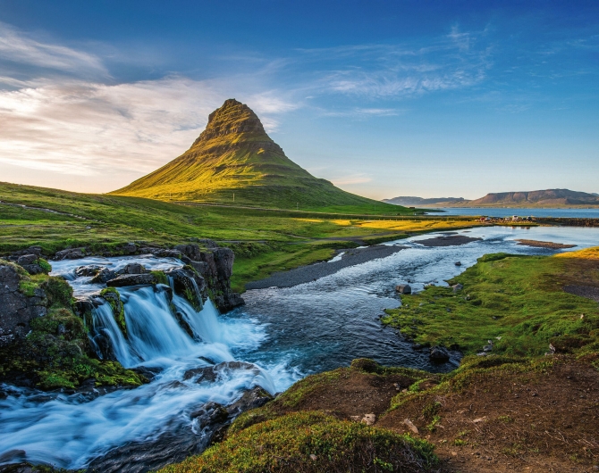 Weitwinkel-Naturaufnahme in Island bei Tageslicht. Im linken Vordergrund stürzt ein kleiner, mehrstufiger Wasserfall über moosbewachsene, dunkle Felsen. Das schäumende, weiße Wasser fließt in einem weiten Bogen als blauer Fluss durch eine saftig grüne, hügelige Landschaft nach rechts hinten weg. Im Bildzentrum erhebt sich im Hintergrund der markante, pyramiden- oder kegelförmige Berg Kirkjufell, dessen linke Flanke von der tief stehenden Sonne hellgrün erleuchtet wird. Rechts im Hintergrund ist ein ruhiger Fjord zu sehen, der von flachen Bergen gesäumt wird. Der Himmel ist strahlend blau mit einigen lockeren, weißen Schleierwolken. Die Lichtstimmung ist klar und spätnachmittags- oder morgendlich warm.