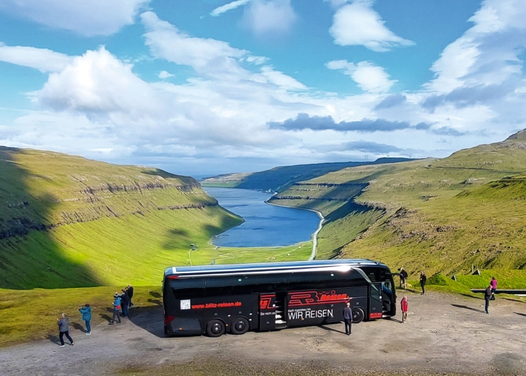 Ein moderner, dreiachsiger Reisebus in Schwarz mit roter und weißer Aufschrift „WIR REISEN“ steht auf einem Schotterparkplatz auf einer Anhöhe. Mehrere Fahrgäste stehen um den Bus herum, einige fotografieren die Landschaft. Im Hintergrund erstreckt sich ein tiefer, blauer Fjord, der von saftig grünen, sanft abfallenden Bergen und Hügeln gesäumt wird. Der Himmel darüber ist hellblau und mit kontrastreichen, weißen Wolken bedeckt, die Schatten auf die Bergflanken werfen.