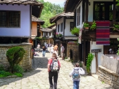 Fotografie einer historischen Gasse in einem Freilichtmuseum unter sommerlichem Tageslicht. Der leicht ansteigende Weg ist mit unebenen, hellen Natursteinplatten gepflastert. Die Gasse wird von traditionellen, mehrstöckigen Fachwerkhäusern mit weißen und ockerfarbenen Fassaden gesäumt. Die Gebäude besitzen markante, weit vorkragende Holzkonstruktionen im Obergeschoss und sind mit dunklen, flachen Steinplatten (Schiefer) gedeckt. Am rechten Haus hängt ein Schild in kyrillischer Schrift mit einem Pfeil nach rechts sowie ein traditioneller, rot-schwarz gestreifter Webteppich über der Balkonbrüstung. Im Vordergrund laufen zwei Jungen mit Rucksäcken von hinten gesehen die Gasse hinauf. Weiter hinten im mittleren Bildbereich verteilen sich mehrere Touristen, die spazieren gehen oder am Wegrand stehen. Der Hintergrund öffnet sich zu dichtem, grünem Wald.