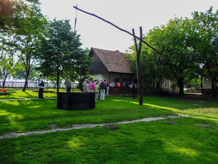 Fotografie eines weitläufigen, grünen Hofgartens unter einem hellen Himmel mit starkem Sonnenschein von rechts oben. Im linken Vordergrund steht die dunkle, hölzerne, runde Einfassung eines Brunnens. Rechts daneben ragt die markante, hölzerne Konstruktion eines traditionellen Ziehbrunnens (Schadoof) empor, dessen langer, schräger Hebelarm weit in die Luft ragt. Im mittleren Hintergrund steht eine Gruppe von etwa 15 älteren Besuchern in sommerlicher Freizeitkleidung auf dem Rasen. Ein Mann links außen und ein Mann rechts der Gruppe tragen dunkle Westen und weiße Hemden, die an traditionelle Festtagskleidung erinnern. Hinter der Gruppe erstreckt sich ein ländliches, weiß getunktes Bauernhaus mit einer überdachten Veranda und einem ziegelroten Satteldach. Das gesamte Areal ist von dichten, grünen Laubbäumen und Rasenflächen umgeben, auf denen sich Schatten und helle Sonnenflecken abwechseln.
