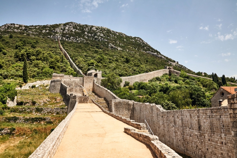 Weitwinkel-Fotografie der historischen Festungsmauer von Ston in Kroatien unter einem leicht bewölkten, hellblauen Himmel. Im Bildvordergrund führt ein breiter, hellbraun gepflasterter Wehrweg geradeaus nach hinten. Er ist auf beiden Seiten von hohen, hellgrauen Mauern aus Natursteinquadern begrenzt. Im Hintergrund teilt und verzweigt sich die Wehranlage an einer kleinen Festungsbastion mit einem Rundbogen-Tor. Von dort aus zieht sich eine schmalere, zinnenbewehrte Steinmauer in steilen Serpentinen und geraden Linien den gesamten, dicht mit grünen Büschen und Bäumen bewachsenen Berghang hinauf bis zum Berggipfel. Rechts am Fuß des Hanges ist das Dach eines Steinhauses mit roten Ziegeln erkennbar. Auf der linken Seite des Weges befinden sich terrassierte Grünflächen.