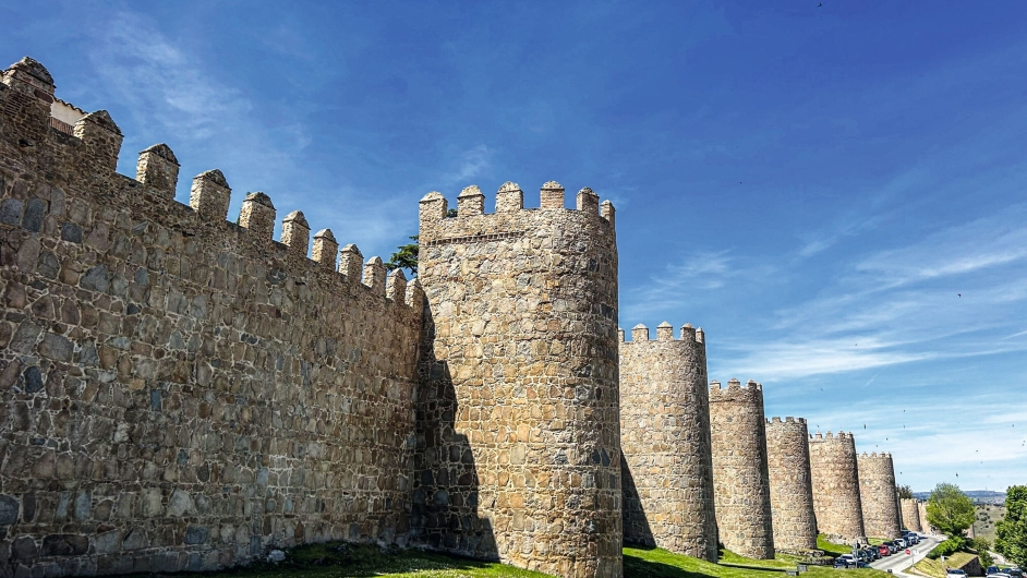Ein querformatiges Foto der monumentalen, mittelalterlichen Stadtmauer von Ávila an einem sonnigen Tag unter einem intensiv blauen, fast wolkenlosen Himmel. Die Aufnahme wurde aus einer niedrigen Perspektive direkt am Fuß der Befestigungsanlage gemacht.Auf der linken Seite ragt die mächtige, hohe Wehrmauer aus grob behauenem, hellbraunem und grauem Naturstein auf, die am oberen Rand lückenlos mit spitzen Steinzinnen besetzt ist. Das Mauerwerk folgt einer leichten Kurve und erstreckt sich in einer langen, rhythmischen Fluchtlinie weit in den rechten Hintergrund hinein. In regelmäßigen Abständen springen massive, halbrunde Festungstürme (Wehrtürme) aus der Wand hervor, die ebenfalls mit Zinnen gekrönt sind. Der vorderste, zentrale Turm wirft einen harten Schatten auf den linken Mauerabschnitt.Am unteren Bildrand zieht sich ein schmaler Streifen eines saftig grünen Rasenhangs entlang, an dem die Fundamente der meterdicken Steinmauer verankert sind. Ganz im rechten Hintergrund ist im Anschnitt eine Straße mit geparkten Autos und etwas modernerer Vegetation zu erkennen, was den Kontrast zwischen historischer Befestigung und heutiger Infrastruktur verdeutlicht. Das helle Sonnenlicht hebt die raue Oberflächenstruktur und die Erdtöne der historischen Granitsteine plastisch hervor.