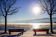 Zwei Bänke im Schnee mit Bäumen, Blick auf einen See bei Sonnenaufgang.
