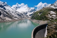 Stausee in den Alpen mit schneebedeckten Bergen und Staumauer im Vordergrund.