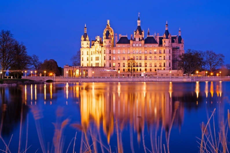 Beleuchtetes Schloss am Abend, reflektiert im ruhigen Wasser.