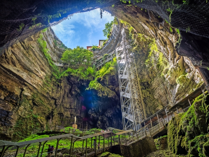 Blick aus einer Höhle nach oben, mit Pflanzen und blauem Himmel sichtbar.