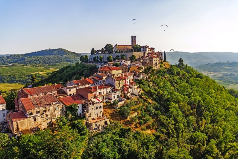 Blick auf eine historische Stadt auf einem Hügel mit Paragleitern am Himmel.
