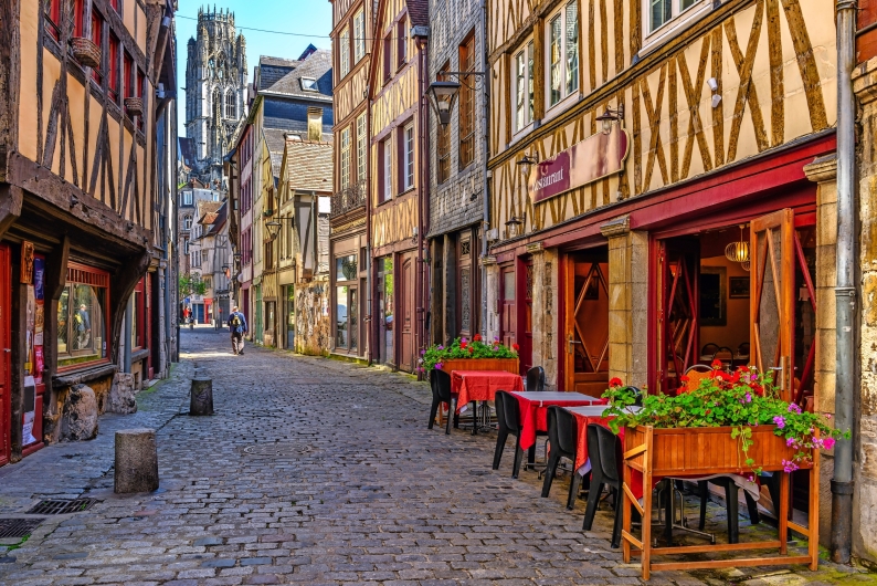 Gepflasterte Altstadtstraße mit Fachwerkhäusern und Straßencafé, Radfahrer im Hintergrund.