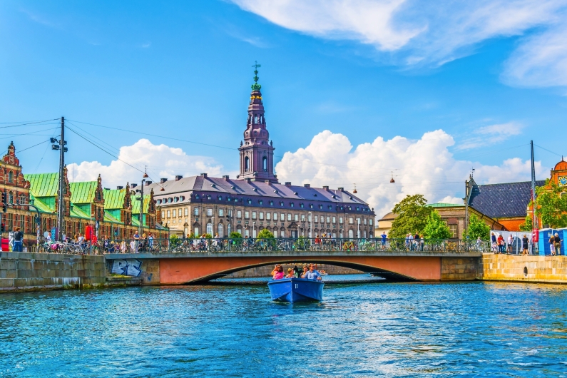 Menschen in einem blauen Boot auf einem Kanal vor historischem Gebäude an einem sonnigen Tag.