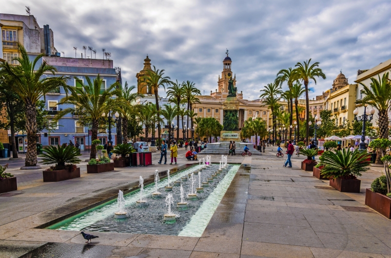 Platz mit Springbrunnen und Palmen, umgeben von historischen Gebäuden.